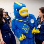 two nursing students in scrubs laugh at a Jaybird mascot with a stethoscope around its neck